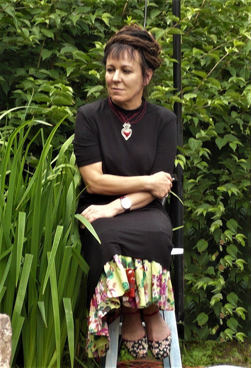 Zdjęcie przedstawia dojrzałą kobietę. Jej twarz jest pociągła, oczy ciemne. Kobieta ma długie włosy upięte wokół głowy. Kobieta ubrana jest w czarną sukienkę z kolorową falbaną na dole. Kobieta siedzi. Trzyma się prawą dłonią za łokieć lewej ręki.