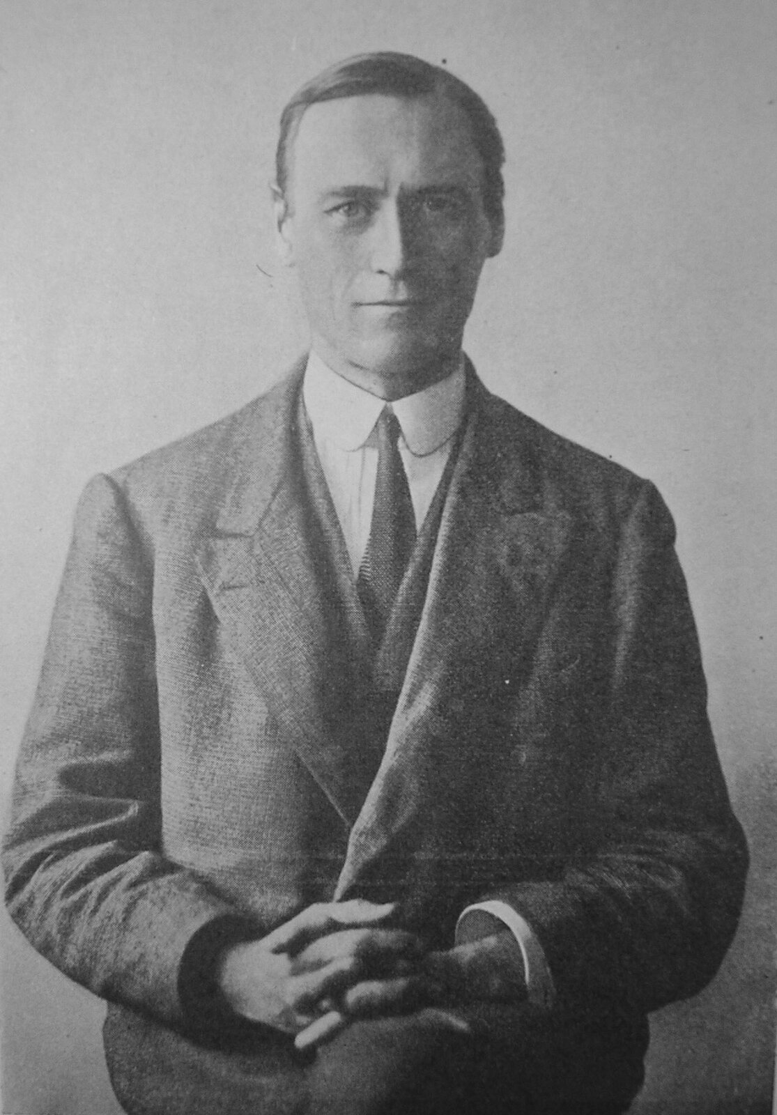 Czarno‑białe zdjęcie przedstawia mężczyznę w pozycji siedzącej, w średnim wieku, z krótkimi, zaczesanymi na bok włosami. Mężczyzna ubrany jest w ciemny garnitur,  białą koszulę i czarny krawat. Dłonie trzyma splecione ze sobą i oparte o kolana.