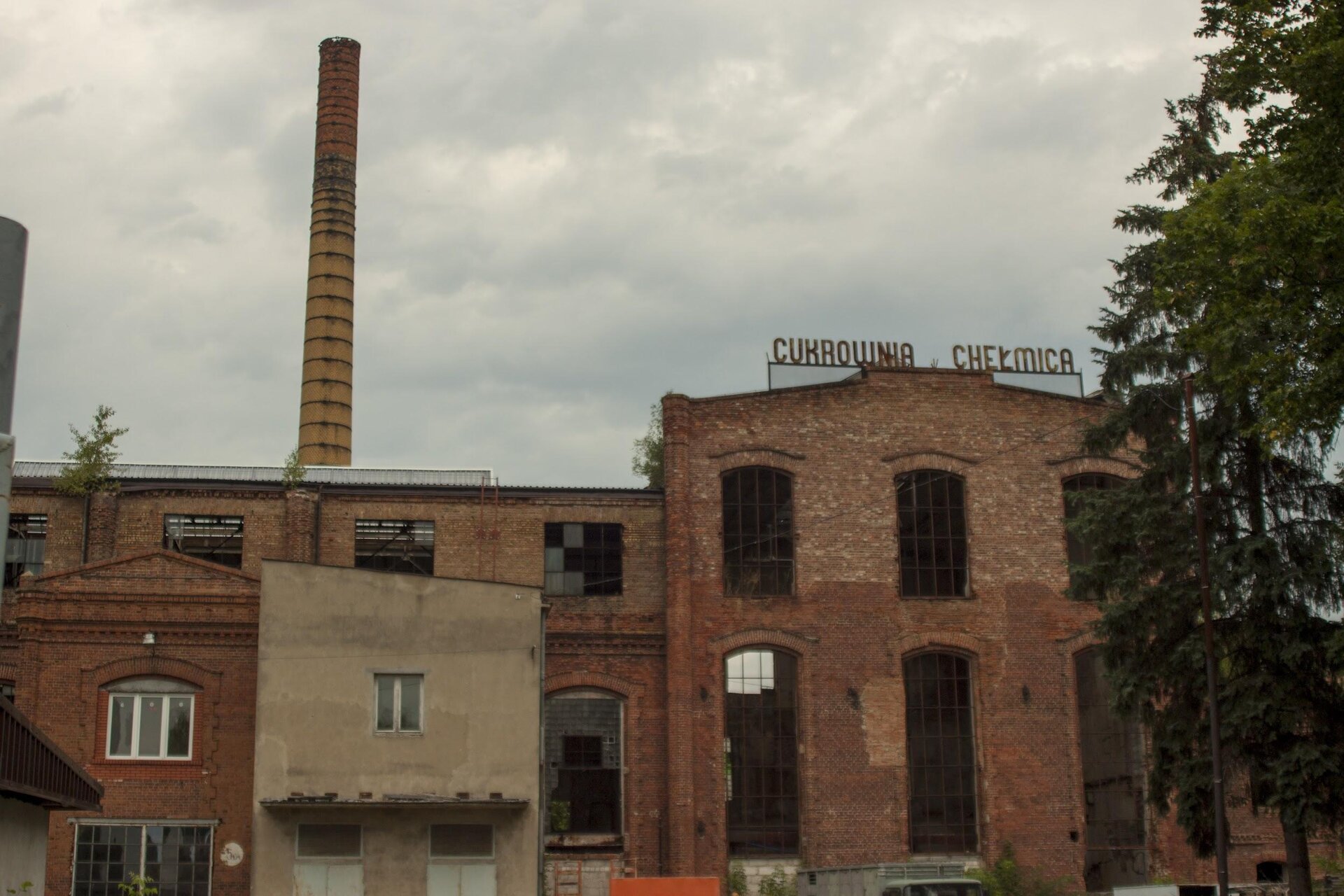 Zdjęcie przedstawia ceglany budynek, u szczytu którego znajduje się napis: "Cukrownia Chełmica". Budynki są poniszczone, a szyby w oknach wybitne. Przed starym budynkiem znajduje się nowszy, niższy, który częściowo pokryty jest kremową farbą. W tle widoczny jest wysoki, pomarańczowy komin. Niebo jest zachmurzone. Zdjęcie zostało wykonane w świetle dziennym.