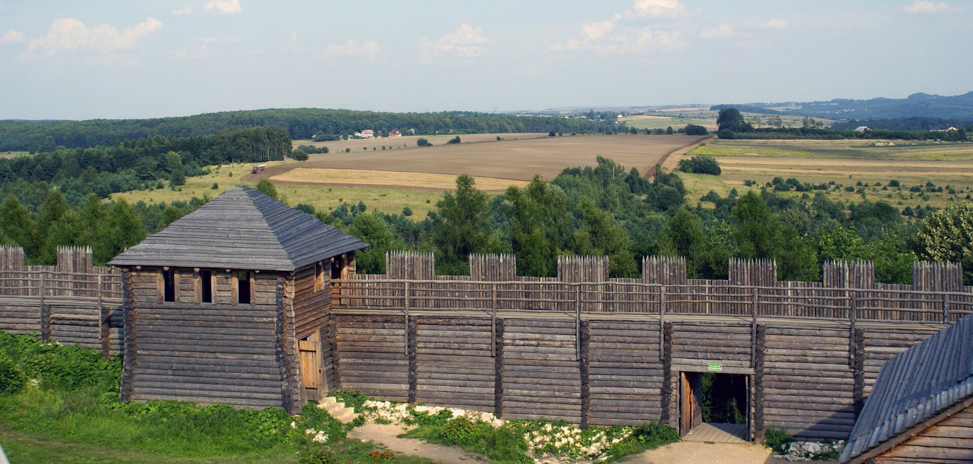 Fotografia  przedstawiająca fragment rekonstrukcji osady i fortyfikacji drewnianej Słowian. Budowlę wykonano z drewnianych bali. W tle widać pola uprawne i lasy, na pierwszym planie stoi drewniana fortyfikacja, do której prowadzi niewielka brama. Fortyfikacja jest kilkumetrowa. Widoczna jest też wieża wartownicza z trzema otworami.