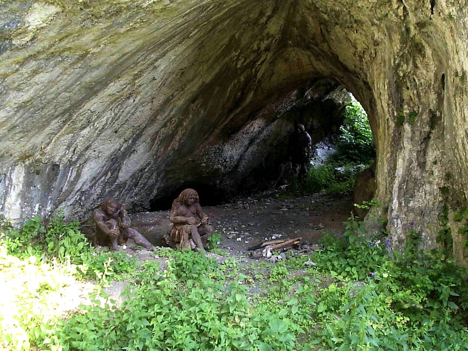 Ilustracja przedstawia rekonstrukcję obozowiska neandertalczyków w Ojcowskim Parku Narodowym. Dwie ciemnoskóre postaci przypominające postaci ludzkie z długimi włosami siedzą przed półokrągłym wejściem do jaskini pod skałą. Przed nimi ułożone są kawałki drewna, na pierwszym planie rosną pokrzywy – niewielkie, zielone rośliny, których łodygę porastają spiczaste liście. 