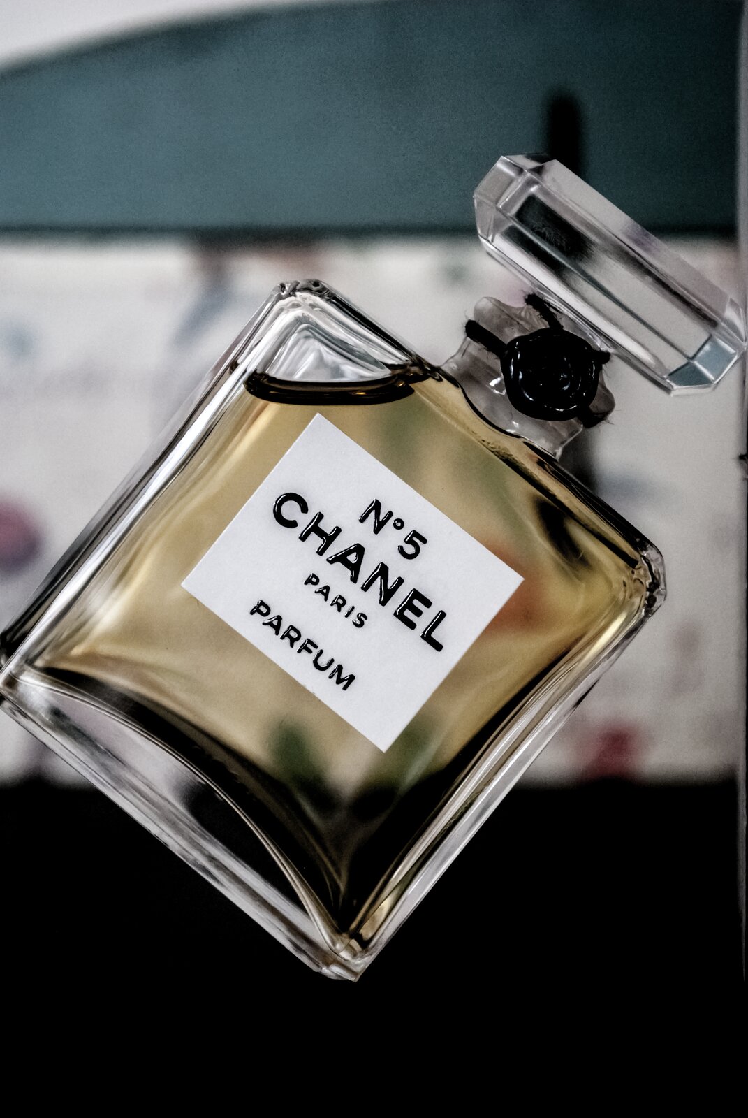 Fotografia przedstawia butelkę damskich perfum Chanel Nr 5,  wprowadzonych na rynek przez kreatorkę mody Coco Chanel,. Butelka jest przechylona, kształtem przypomina kwadrat i jest przezroczysta. W środku widoczny jest złotawy płyn. Zamknięcie perfum również jest przezroczyste. Widać na nim czarny element, wyglądający jak pieczęć. Etykieta to biały prostokąt z przodu butelki z czarnymi napisami „Nr 5 CHANEL, Paris, PARFUM”. 