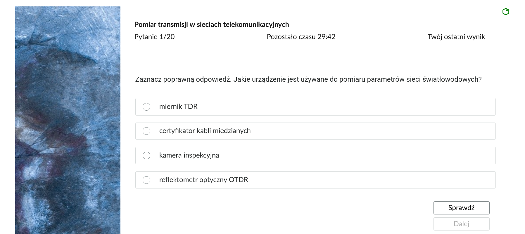 Grafika przedstawia przykładowe zadanie testowe. Ponad poleceniem widać numer pytania, czas, który pozostał na wypełnianie testu, oraz ostatni uzyskany wynik. Pod poleceniem znajdują się możliwe odpowiedzi. W prawym dolnym rogu panelu z zadaniem znajdują się dwa przyciski. Są to "Sprawdź" oraz "Dalej".