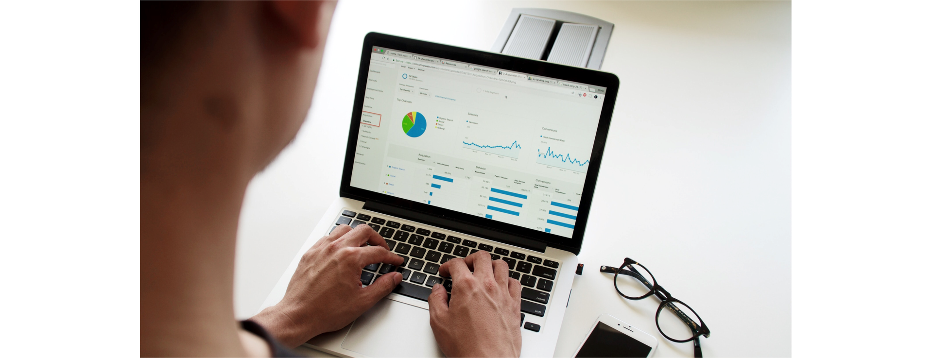 Zdjęcie przedstawia zbliżenie na otwarty laptop. Widoczne są dłonie na klawiaturze laptopa. Na ekranie widoczne są wykresy. Obok laptopa leżą okulary.