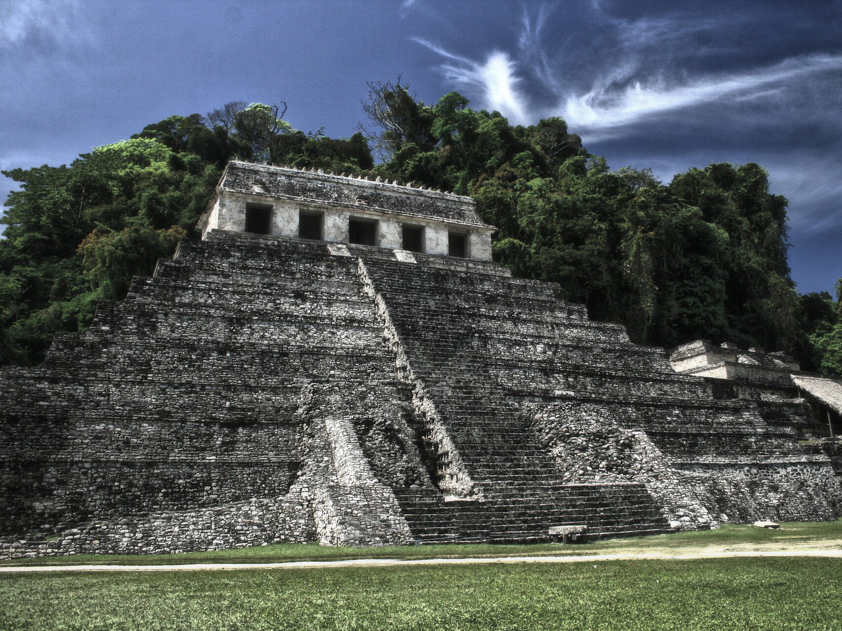 Fotografia barwna przedstawiająca Świątynię Palenque. Ma kształt piramidy, prowadzi do niej wiele schodów. Wokół zasadzone drzewa i bujna roślinność.