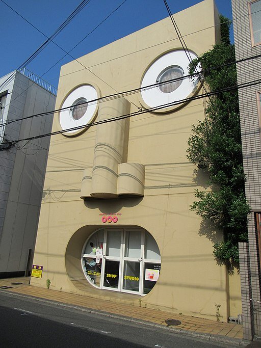 Zdjęcie przedstawia nowoczesny budynek, stojący przy ulicy. Jego front posiada owalne, przeszkolone wejście przypominające otwarte, ludzkie usta. Nad wejściem znajdują się trzy walcowate kształty przypominające ludzki nos. Powyżej nosa są dwa okrągłe okna przypominające ludzkie oczy.