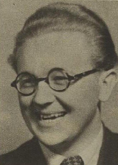 Zdjęcie przedstawia uśmiechniętego mężczyznę. Ma owalną twarz, szeroki nos i okulary w okrągłych oprawkach. Ma zaczesane do tyłu włosy i ubrany jest w garnitur z krawatem.