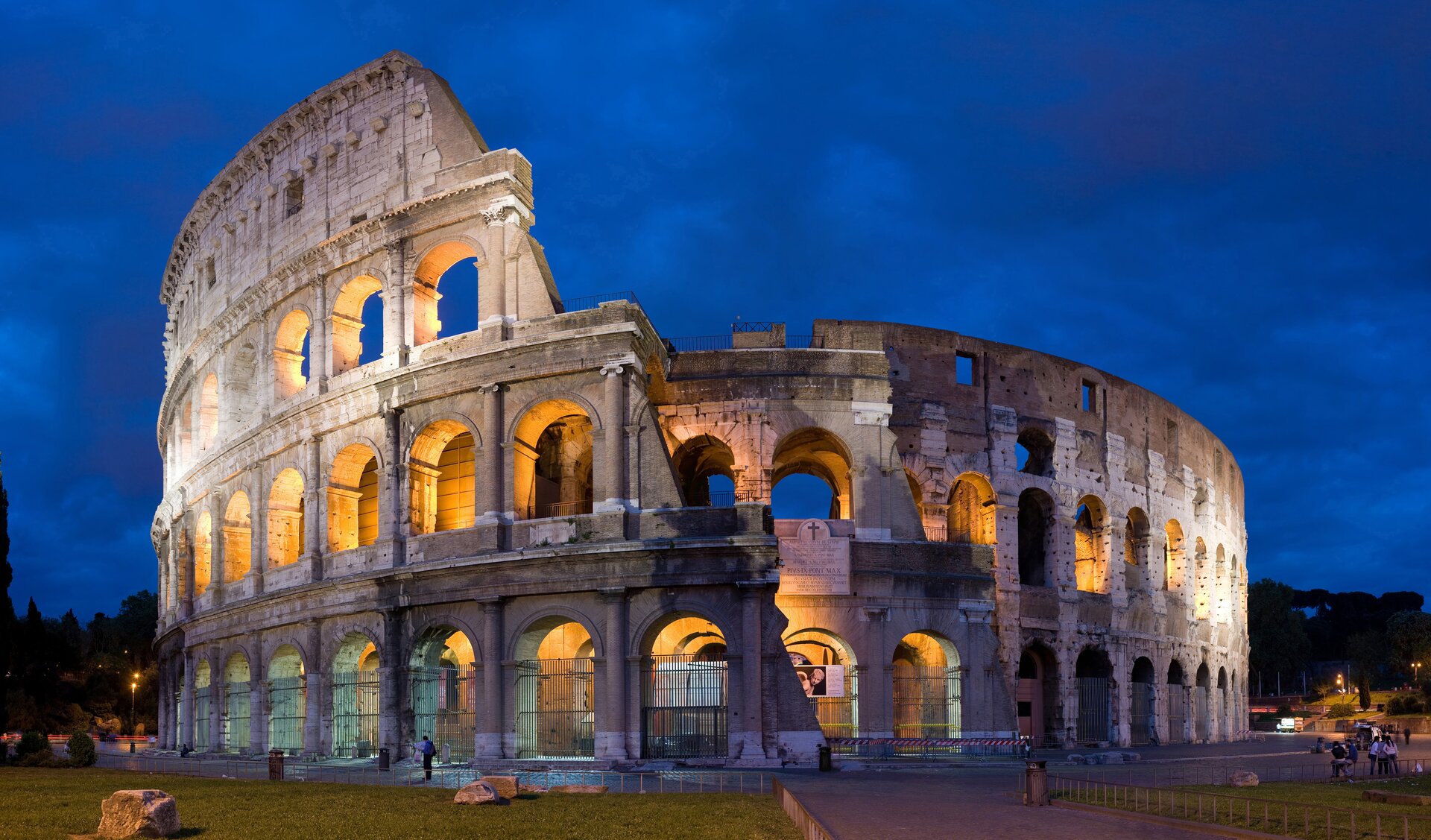 Ilustracja banerowa przedstawia fragment Koloseum, oficjalnie w starożytności Amfiteatr Flawiuszów wzniesiony w latach 70-72 do 80 n.e. przez cesarzy z dynastii Flawiuszów. W kadrze znajduje się górna część budowli, częściowo w ruinie. wszystkie cztery kondygnacje są oddzielone gzymsem. najmniej widoczna jest najniższa i najwyższa kondygnacja. Obiekt przedstawiony jest na tle ciemnego nieba i wewnątrz oświetlony żółtym światłem reflektorów.