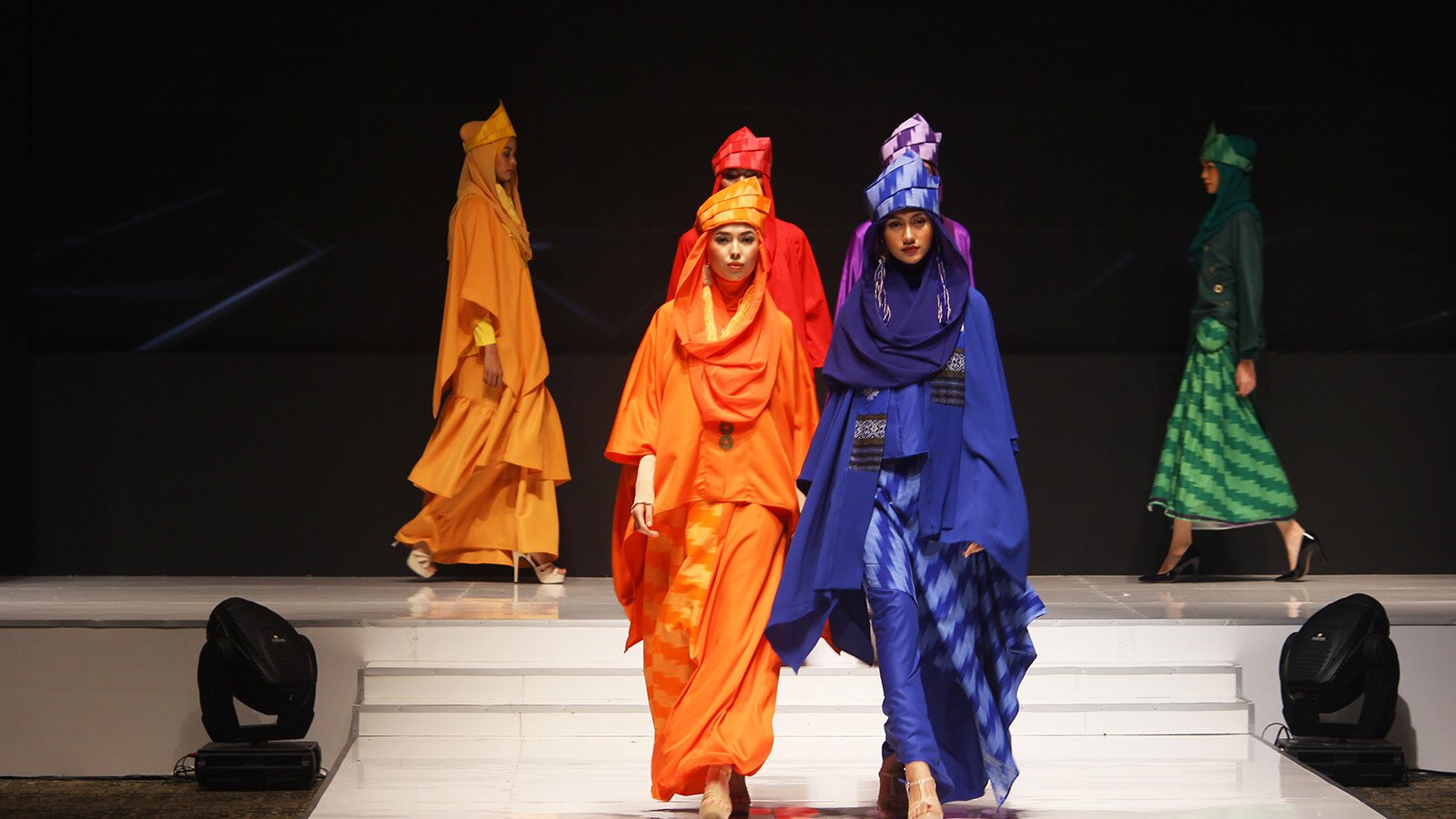 Zdjęcie przedstawia fragment wybiegu, po którym idą obok siebie dwie modelki. Kobiety mają na sobie luźne, kolorowe sukienki do samych kostek, z długim rękawem. Na głowie, obie Panie mają nowoczesne turbany z tego samego materiału co ich suknie.