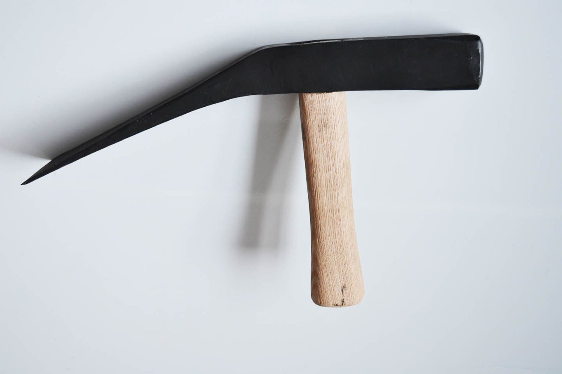 Fotografia przedstawia młotek kamieniarski o czarnym metalowym obuchu i rąbie w kształcie klina. Młotek ma krótki trzonek wykonany z jasnego drewna. Tło stanowi biała ściana, na której widoczny jest cień rzucany przez narzędzie.