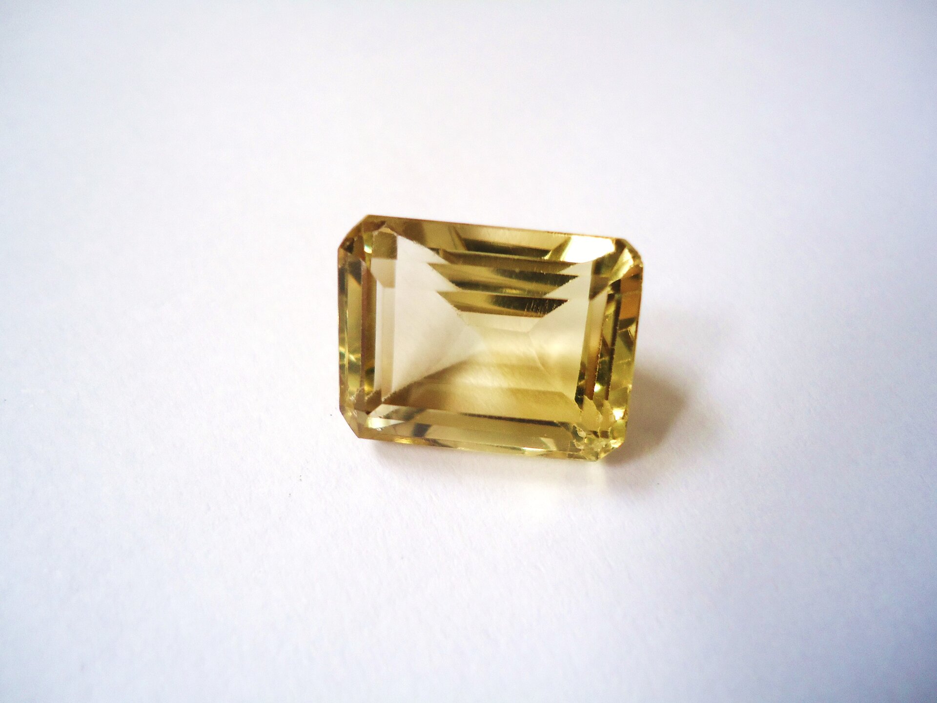 Zdjęcie przedstawia oszlifowany cytryn w formie szlifu szmaragdowego, czyli prostokątnego. Kamień jest jasnożółty i lekko przejrzysty, leży na białym tle.