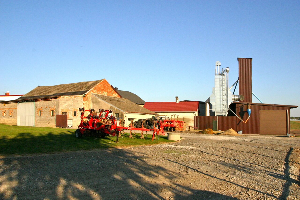 Zdjęcie przedstawia budynki gospodarstwa. Na pierwszym planie jest zaorane pole i maszyna rolnicza.