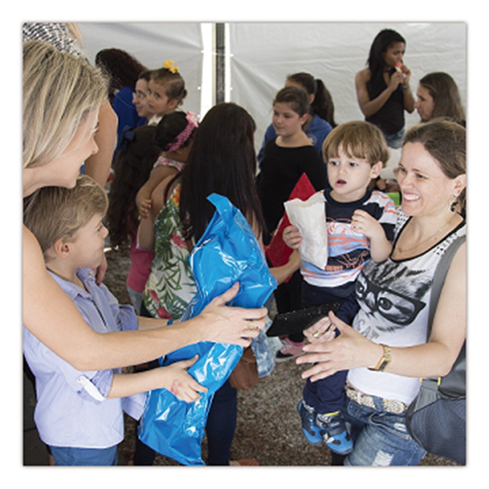 Na zdjęciu znajduje się grupa ludzi w dużym namiocie. Kobieta z synkiem podają torbę innej kobiecie, która trzyma na rękach dziecko. 
