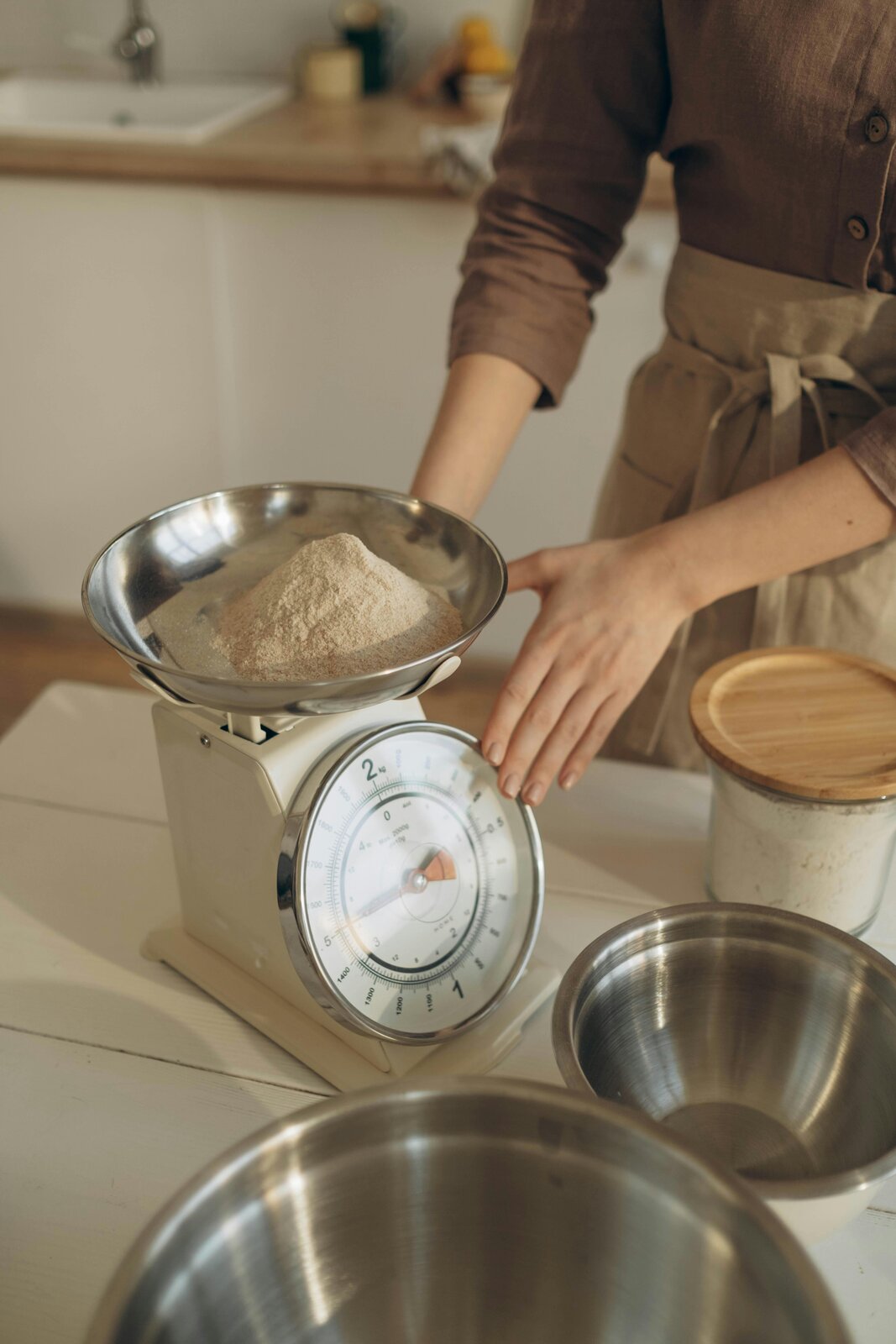 Trzecie zdjęcie jest podpisane Waga. Na zdjęciu widoczna jest klasyczna waga kuchenna. Ma białą obudowę i wyposażona jest w okrągłą tarczę z podziałką i wskazówką do odczytu masy. Na górnej, metalowej misce wagi znajduje się mąka. Obok wagi, na jasnym blacie kuchennym, umieszczone są inne metalowe naczynia.