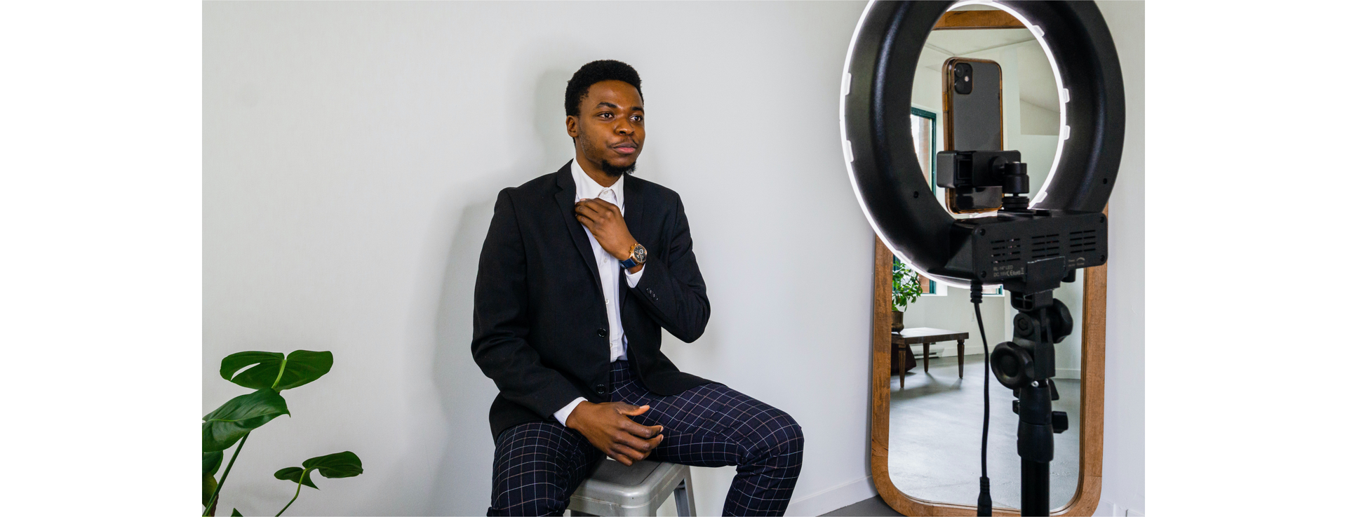 Zdjęcie przedstawia czarnoskórego chłopaka w garniturze. Chłopak siedzi na stołku przed telefonem komórkowym znajdującym się na statywie. Na statywie znajduje się także okrągła lampa oświetlająca mężczyznę.