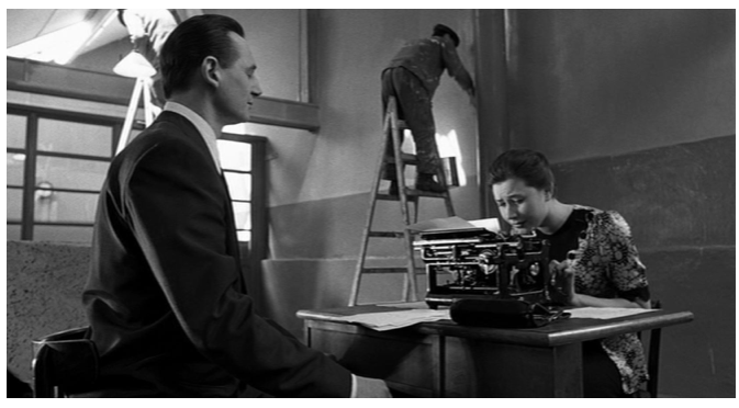 Zdjęcie przedstawia wnętrze fabryki. Robotnicy malują ściany. W sali stoi biurko, przy którym siedzi przy maszynie do pisania młoda kobieta o ciemnych włosach. Naprzeciw niej siedzi Schindler. Kobieta pochyla się nad maszyną do pisania.