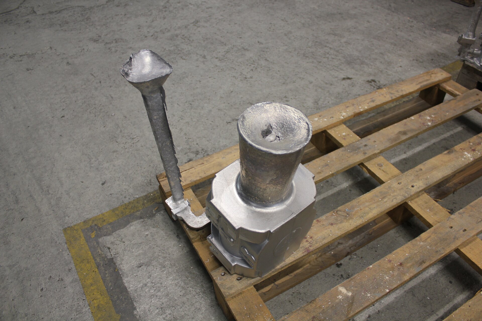 Ilustracja przedstawia odlew metaliczny, ustawiony na palecie w zakładzie przemysłowym. Odlew składa się z odwzorowanego układu wlewowego oraz przelewu.