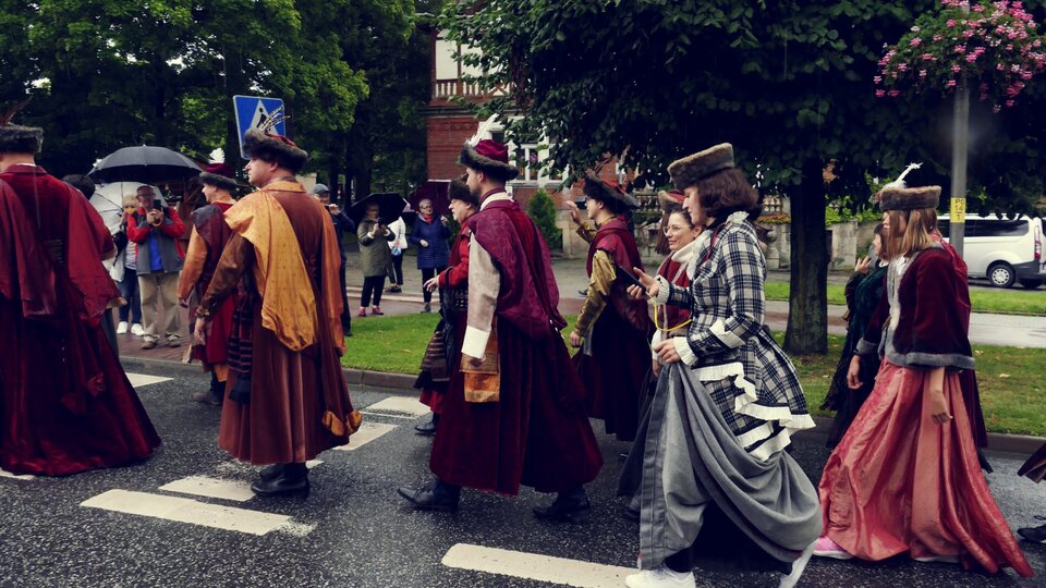 Na zdjęciu znajduje się grupa ludzi idących ulicą. Mężczyźni ubrani są w długie bordowe płaszcze. Kobiety noszą długie suknie. Wszyscy mają na głowach czapki obszyte futrem. W tle zdjęcia znajdują się ludzie robiący zdjęcia, drzewa oraz domy.