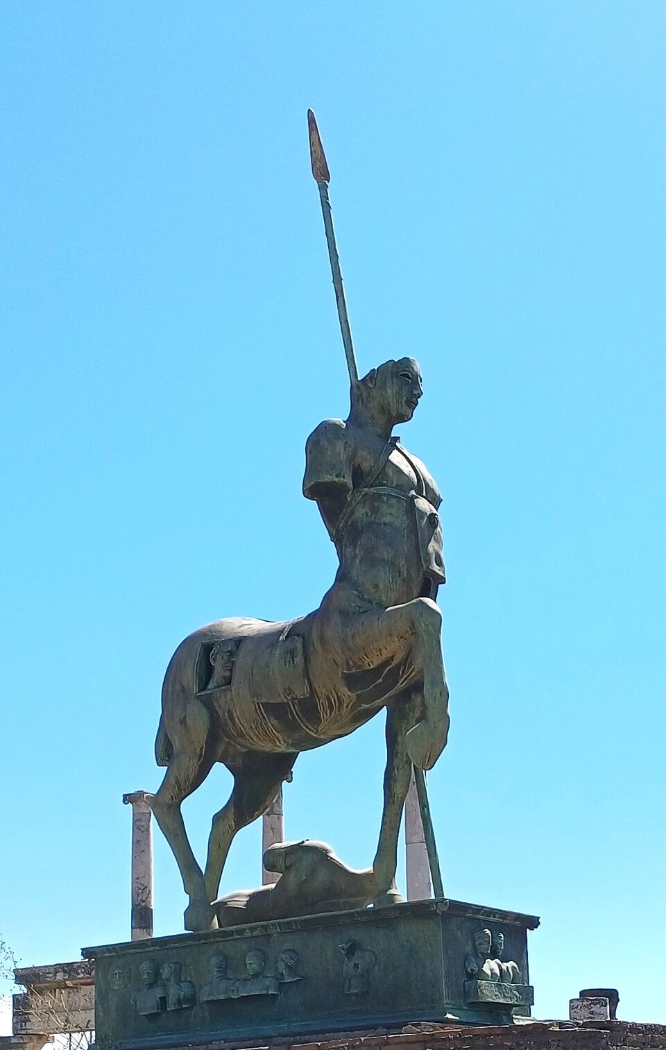 Fotografia przedstawia monumentalną rzeźbę z brązu. Na prostokątnym podeście udekorowanym płaskorzeźbami popiersi, stoi centaur - pół człowiek, pół koń. W jego brzuchu wycięty jest prostokątny otwór, z którego wyłania się męska głowa. Centaur ma zgiętą w kolanie i uniesioną lekko do przodu przednią nogę, oderwane ręce na wysokości ramion oraz głowę od czoła w górę. O bok konia opiera się wysoka włócznia, zaś pod jego kopytami leży umięśniony męski korpus. 