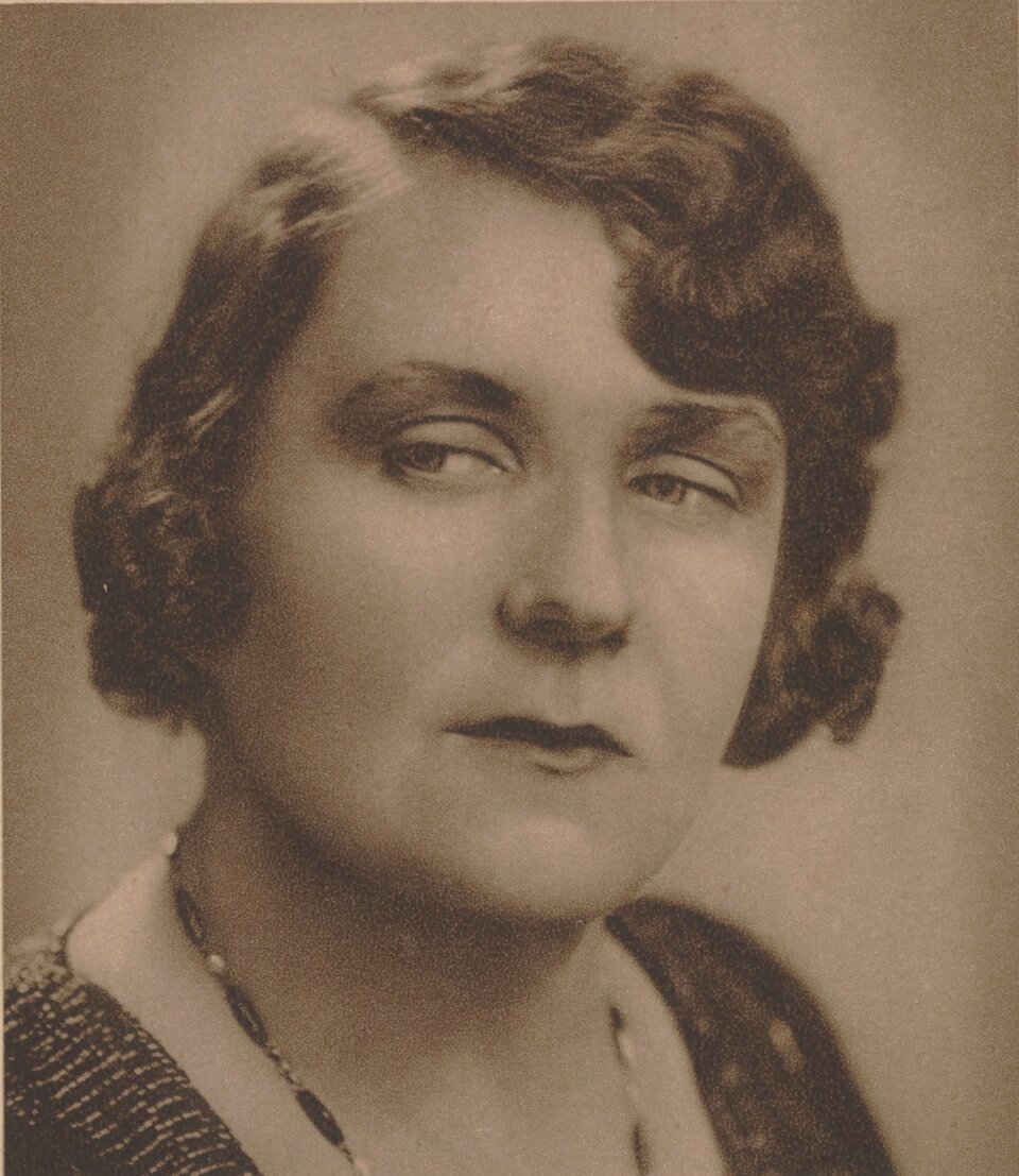 Zdjęcie przedstawia portret dojrzałej kobiety. Ma ona niedługie, pofalowane włosy. Są one starannie ułożone wokół twarzy. Kobieta ma okrągłą twarz. Jej oczy są duże, okalają je od góry wyraziste brwi. Kobieta spogląda w swoją prawą stronę. Ma poważny wyraz twarzy. Na jej szyi wisi delikatny naszyjnik.