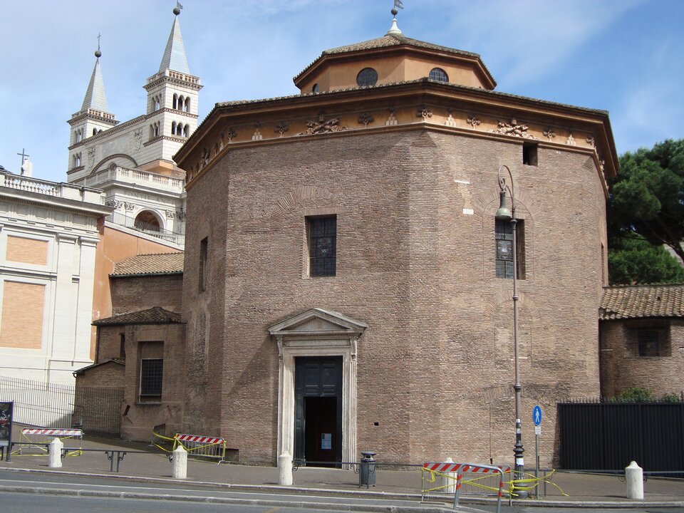 Zewnętrzny widok ośmiobocznego baptysterium z cegły: masywna bryła z małymi, prostokątnymi oknami i kamiennym portalem wejściowym zwieńczonym trójkątnym naczółkiem. W tle widoczna jest jasna fasada i dwie wieże pobliskiej bazyliki, a przed budynkiem biegnie ulica z chodnikiem, barierkami i latarnią; całość oświetlona jest dziennym światłem pod błękitnym niebem.