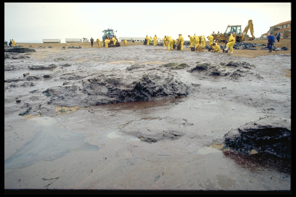 Fotografia przedstawia skutki wycieku ropy po katastrofie tankowca Erika. Na pierwszym planie widać rozległe, ciemne płaty mazutu pokrywające prawie całą powierzchnię plaży oraz przybrzeżne skały. Zanieczyszczenie jest zwarte i bardzo wyraźne, przez co naturalny kolor piasku prawie nie jest widoczny. W dalszej części kadru znajdują się ludzie pracujący przy oczyszczaniu brzegu oraz sprzęt wykorzystywany do usuwania ropy. Zdjęcie pokazuje dużą skalę skażenia wybrzeża i trudność w likwidacji skutków katastrofy.
