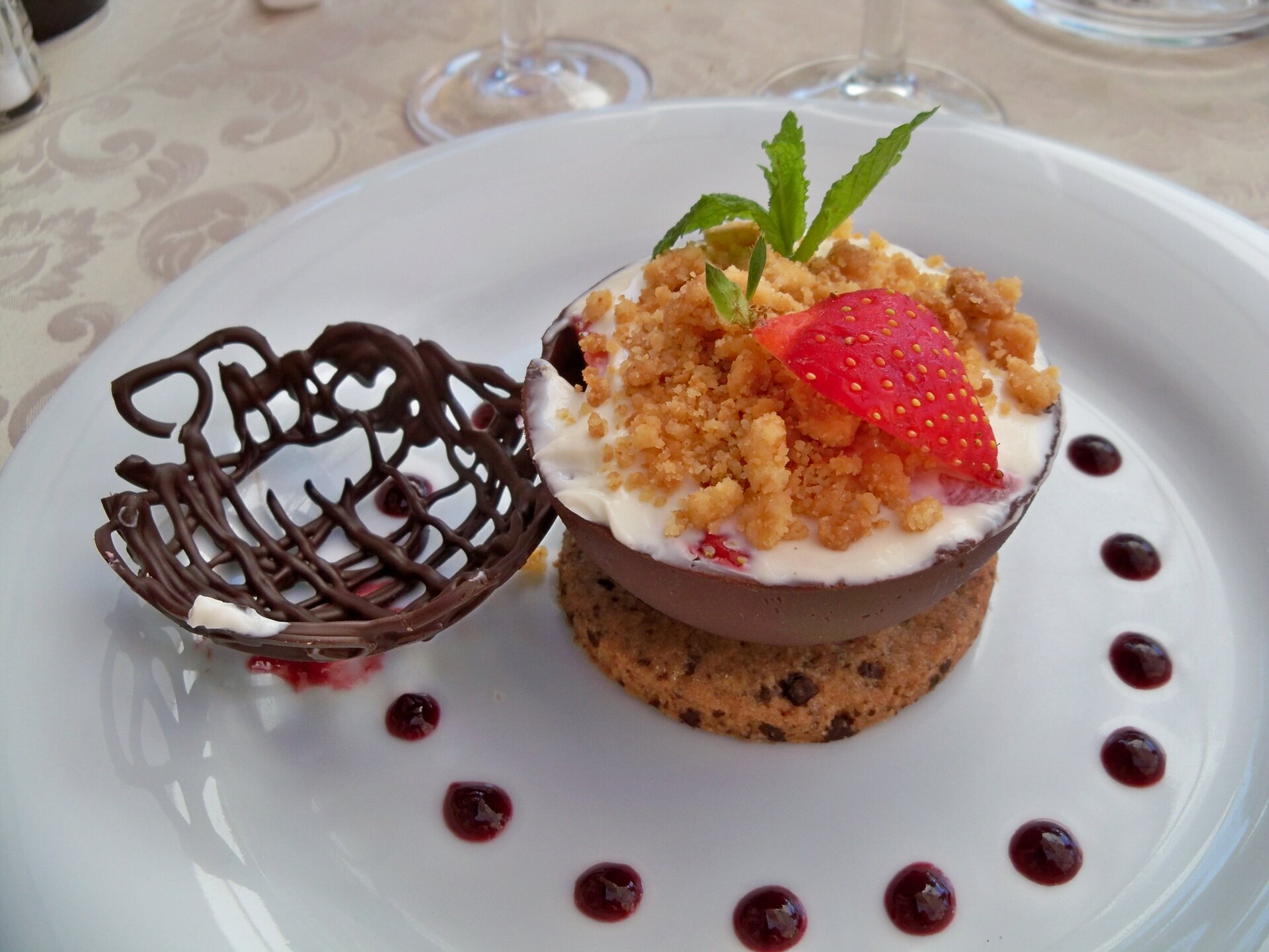 Zdjęcie przedstawia deser w miseczce. Na górze deseru jest kruszonka, a na niej kawałek truskawki. Deser stoi na talerzu ozdobionym dodatkami- czekoladą i sosem truskawkowym.  