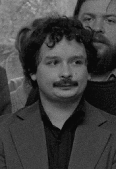 Fotografia przedstawiająca mężczyznę w sile wieku. Postać ukazana jest od piersi w górę, na wprost nas. Mężczyzna ma okrągłą twarz, gładkie czoło, wyraźnie zarysowane brwi, małe oczy, prosty nos, gęste wąsy oraz znamię na lewym policzku. Włosy ma gęste, falujące. Ubrany jest w koszulę i marynarkę. W tle stają inne osoby. 