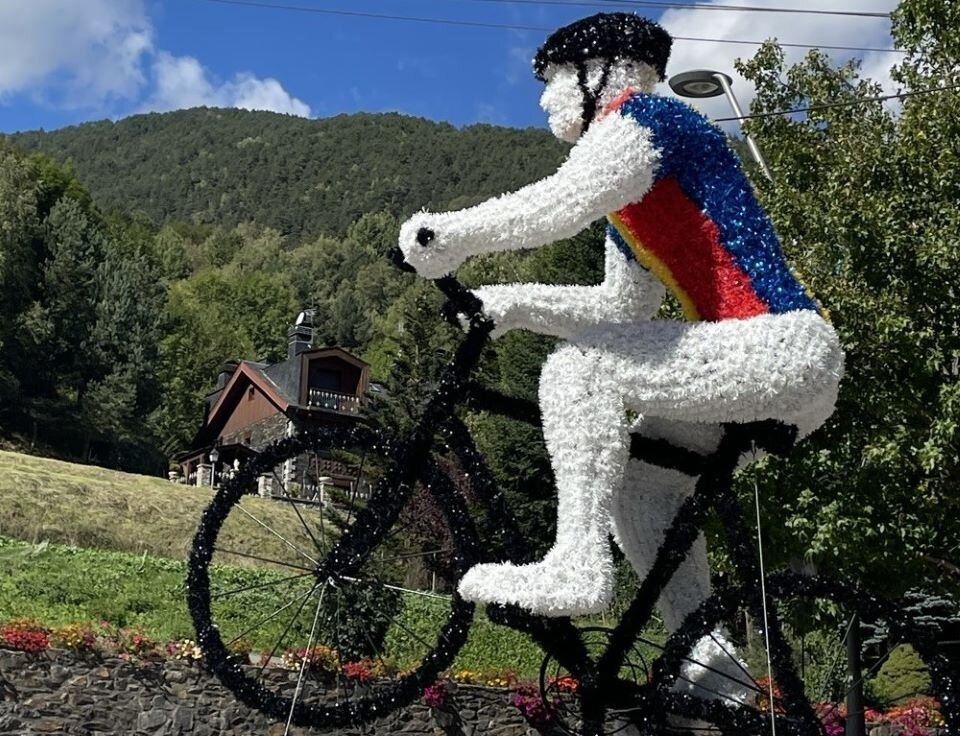 Ilustracja przedstawia rzeźbę rowerzysty. Rzeźba jest wykonana z tworzywa sztucznego i jest ustawiona w przestrzeni na tle trawnika, drzew oraz gór w oddali. Figura rowerzysty jest sfotografowana z rpofilu.