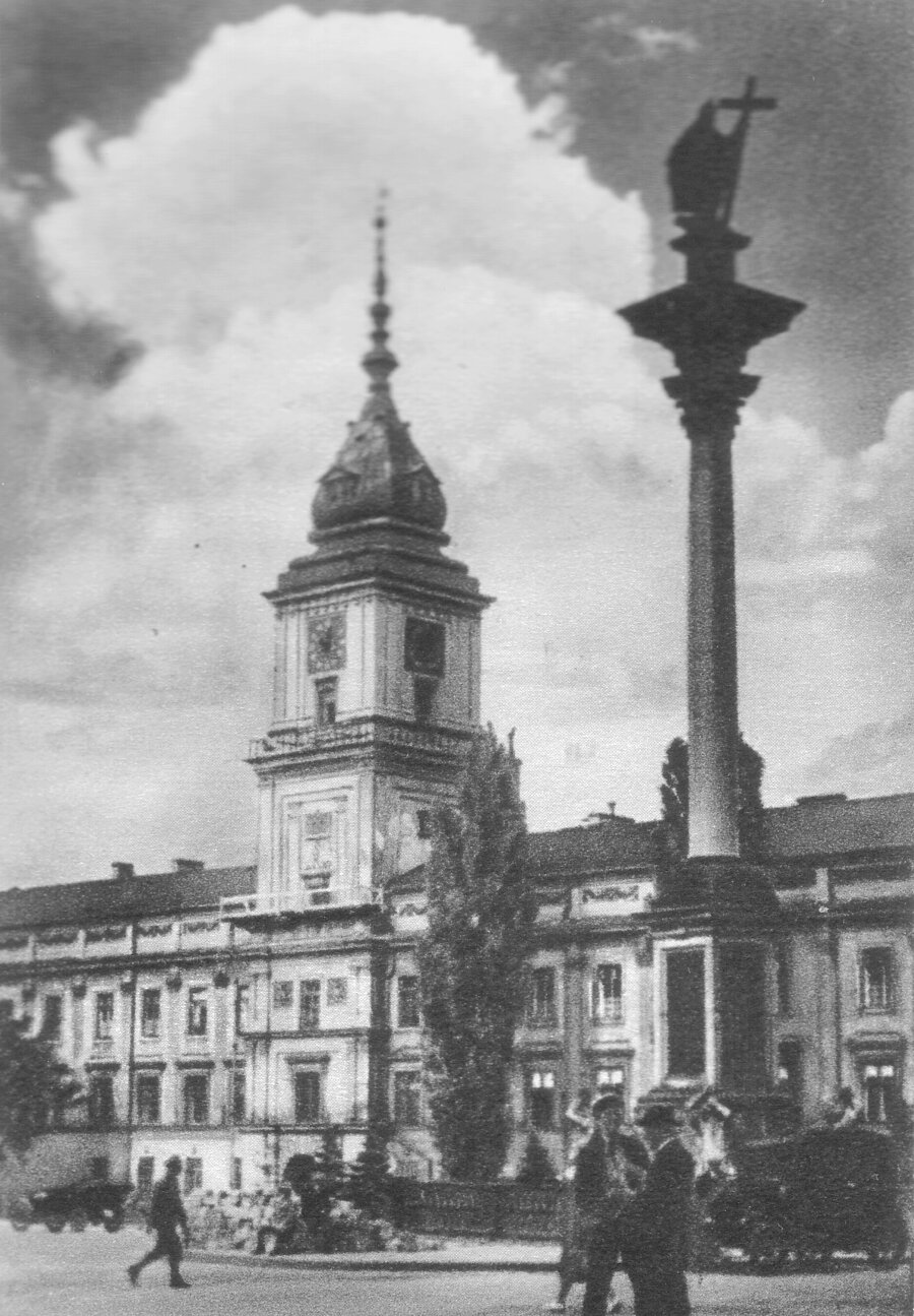 Kliknij, aby powiększyć. Na czarno‑białym zdjęciu znajduje się fasada Zamku Królewskiego w Warszawie i ulica. Zamek wyróżnia się wieżą, która góruje nad dachem. Zamek stoi przy placu, w którego centrum stoi kolumna Zygmunta. Wokół kolumny wiedzie ulica. Na ulicy stoi dwóch ludzi, inny idzie ulicą. Jadą dwa samochody.