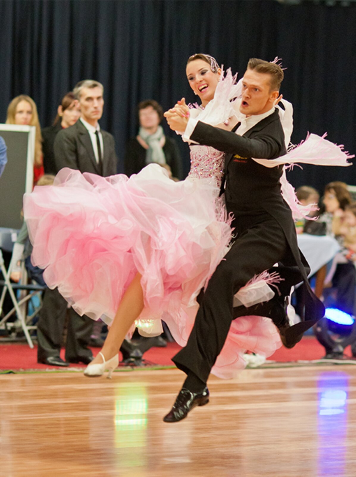 Ilustracja przedstawia fotografię pary tańczącej Foxtrot na Sali Balowej w Huntington. Kobieta ma długą, rozkloszowaną, różową suknię. Góra sukni jest wyszywana różowymi koralikami, a dół ma falbanki. Jej paznokcie są pomalowane na kolor różowy. Na stopach ma białe, zakryte buty na niskim obcasie. Jej włosy są ciemne. Mężczyzna ma czarną marynarkę, spodnie, zakryte, wiązane buty oraz białą koszulę i muszkę. Ma krótkie, ciemne włosy. Mężczyzna trzyma lewą dłonią prawą dłoń kobiety. Para jest bokiem i unosi się nad jasnobrązowym parkietem. W tle znajduje się kilka innych osób. Tło jest rozmyte, niewyraźne. 