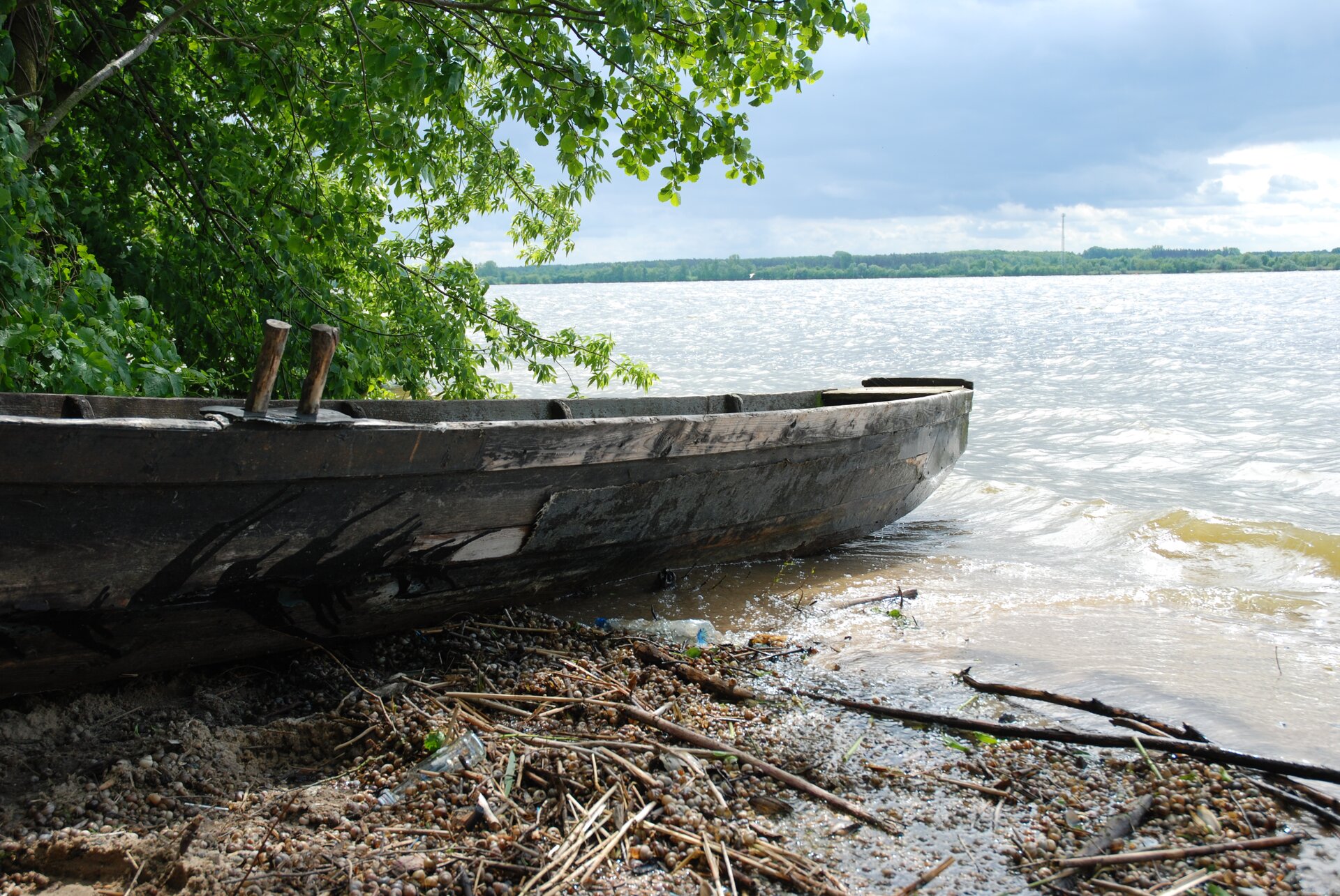 Fotografia przedstawia łódkę na brzegu rzeki. Jest wykonana z drewna, ma długość kilku metrów i szerokość około metra. Na drugim planie szeroka rzeka a za nią zadrzewiony brzeg.