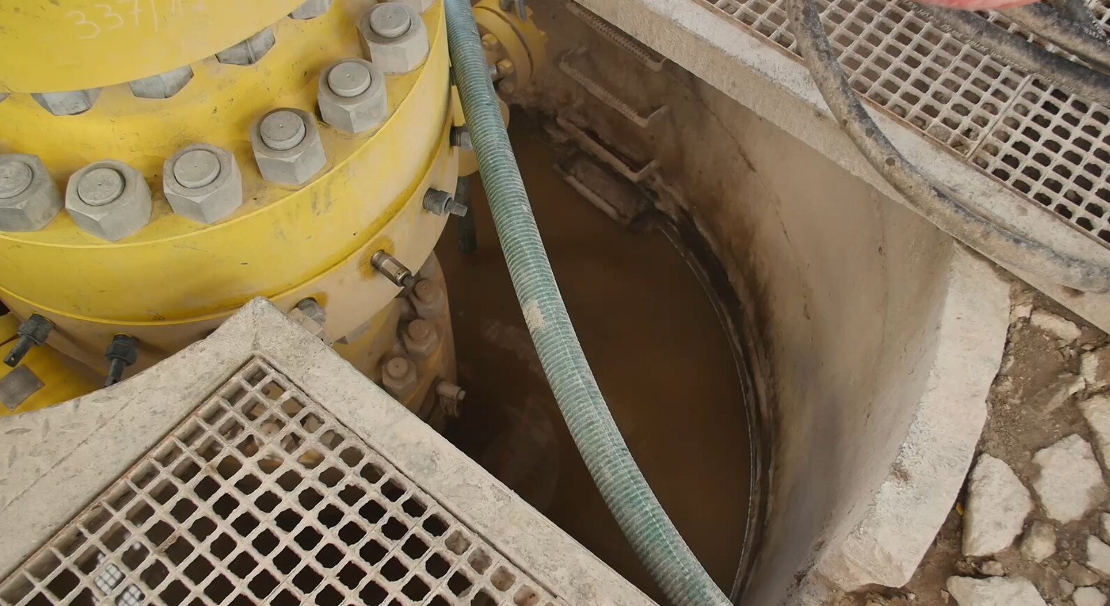 Na zdjęciu widoczna jest więźba rurowa z góry. Więźby rurowe służą do podwieszania rur okładzinowych zapuszczonych i zacementowanych w otworze wiertniczym oraz do instalowania na nich głowic przeciwerupcyjnych. Po zakończeniu wiercenia, gdy otwór przekazywany jest do eksploatacji, na więźbie montuje się głowicę eksploatacyjną. Na zdjęciu widoczny jest otwór. W jego środku znajduje się więźba, a na niej zamontowana jest głowica. W otworze znajduje się płyn. Obok głowicy widoczne są różne przewody.