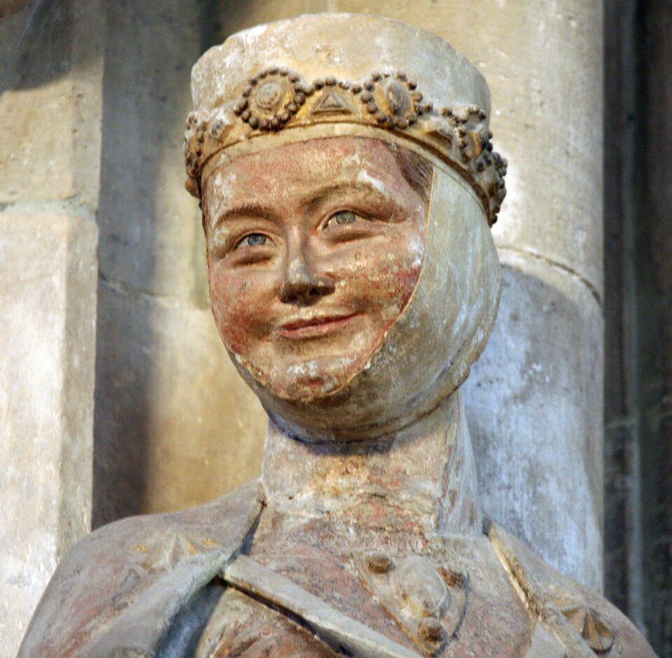 Fotografia przedstawia kamienny posąg kobiety. Widoczne jest tylko jej popiersie. Kobieta jest ubrana w suknię i ma narzucony na ramiona płaszcz. Pod szyją ma chustę, którą ma okręconą głowę przez podbródek, aż do czubka głowy. Nakrycie to zakrywa całkowicie jej włosy. Na chustę nasuniętą ma koronę. Kobieta patrzy przed siebie i uśmiecha się. Ma okrągłą twarz, pucułowate policzki, wąskie usta, uśmiechając się, zwężają jej się oczy. 