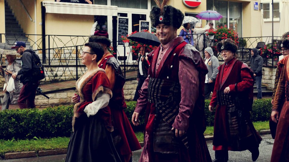 Na zdjęciu znajduje się grupa ludzi idących ulicą. Mężczyźni ubrani są w długie bordowe płaszcze. Kobiety noszą długie suknie. Wszyscy mają na głowach czapki obszyte futrem. W tle zdjęcia znajdują się przechodnie oraz budynki.