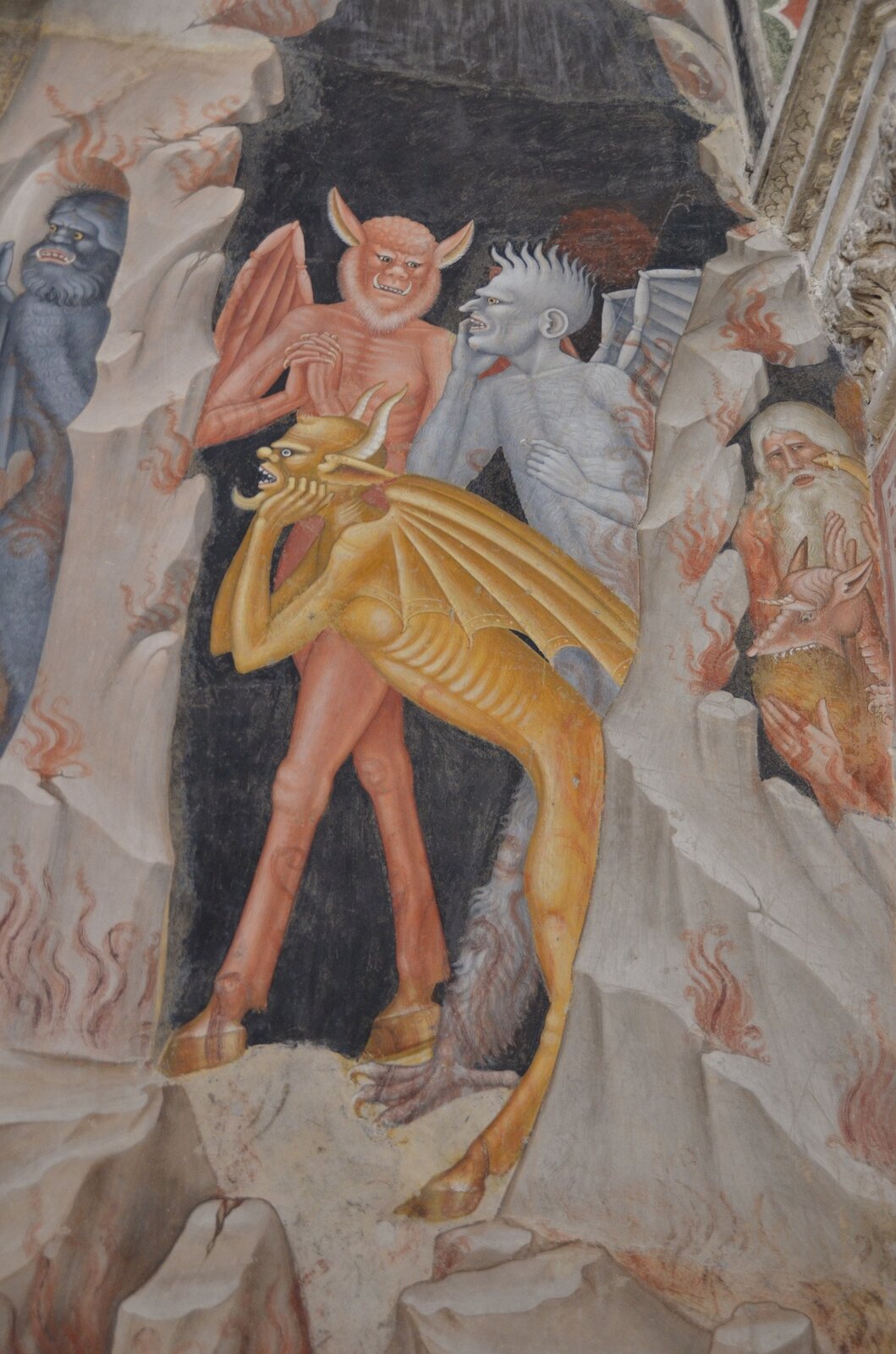 Obraz. Otwarte wejście do jaskini. Kilka postaci diabłów na jej tle. Jeden koloru żółtego, z kopytami, rogami i skrzydłami. Trzyma się rękoma pod brodę, jakby myślał. Dwa pozostałe diabły rozmawiają. Jeden jest koloru pomarańczowego, ma twarz nietoperza, wystające kły, zadarte uszy i skrzydła. Ręce ma jakby złożone do modlitwy. Drugi koloru sinego z wielką, owłosioną, szponiastą łapą, skrzydłami i brzydką twarzą z długim, cienkim nosem. Po lewej w załomku skały widoczna jest jakaś postać  koloru niebieskiego, ze skrzydłami i owłosioną twarzą. Po prawej, w drugim załamaniu skał znajduje się postać świętego. Ma on długie siwe włosy i długą brodę, zamyśloną twarz, ręce składa do modlitwy. Jakieś dziwne stworzenie jakby wgryzało się w jego ciało.