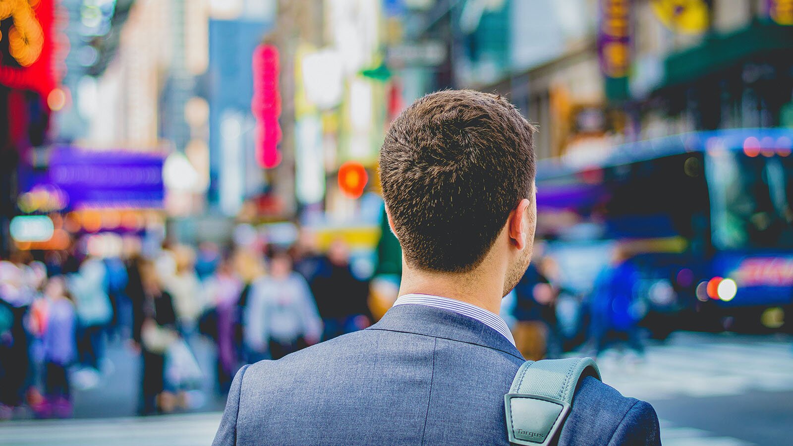 Zdjęcie przedstawia ramiona i tył głowy mężczyzny stojącego na zatłoczonej ulicy. Na jego prawym ramieniu widzimy fragment paska od torby.