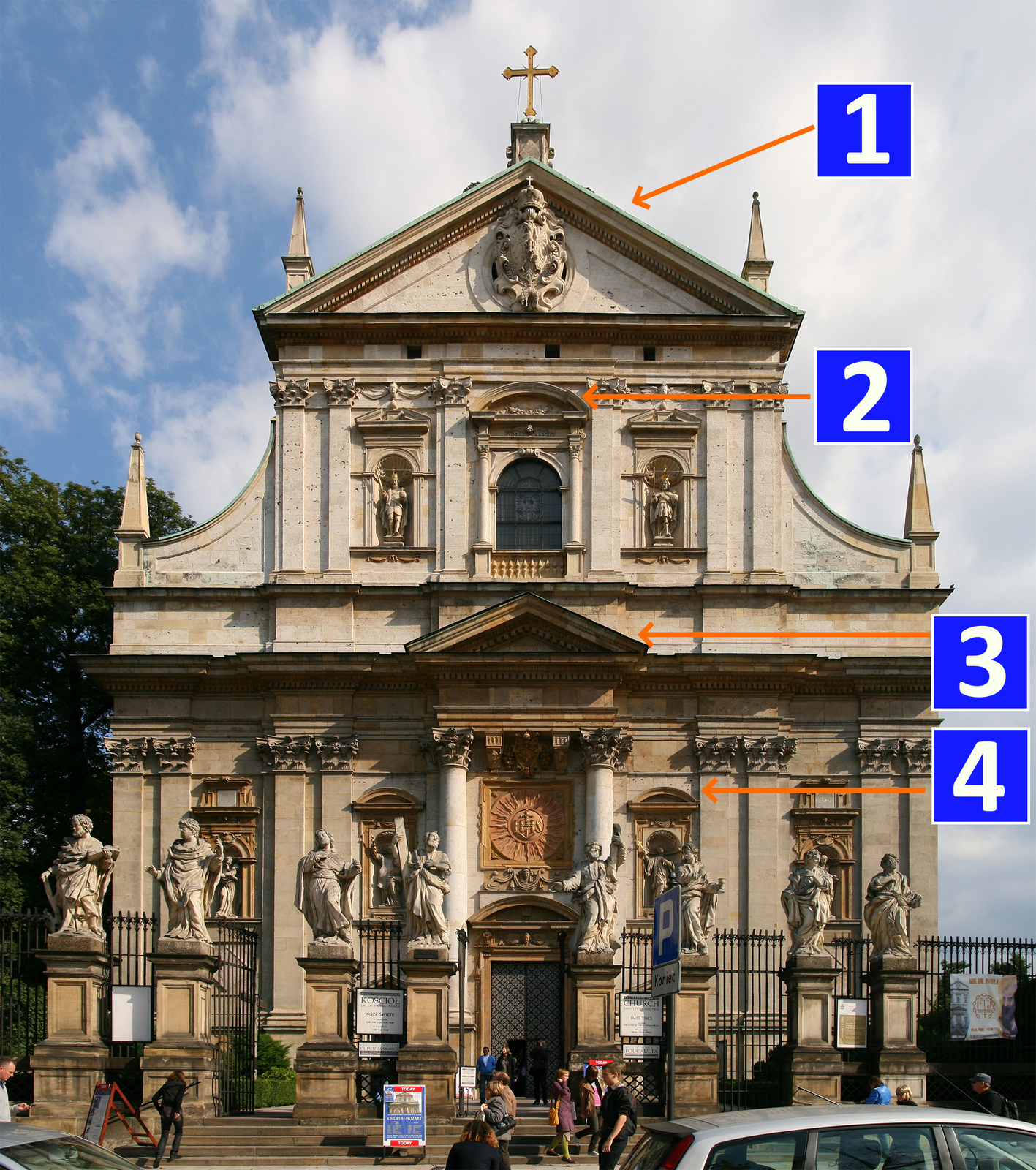 Fotografia przedstawia budynek o bogato zdobionej elewacji w kolorze piasku. Budynek ten posiada duże drzwi wejściowe, figury przedstawiające postaci świętych umieszczone w niszach, dwie kolumny podtrzymujące portyk, balkon obok którego znajdują się kolejne figury świętych. Szczyt zdobi naczółek w kształcie trójkąta na którym znajduje się herb w kształcie pionowej elipsy z której wystają rzeźbione liście, ornamenty, orzeł oraz korona z krzyżem.