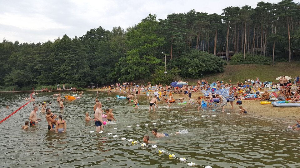 Na zdjęciu jest plaża nad jeziorem. Za plażą jest las. Na plaży są liczni ludzie w strojach kąpielowych, w wodzie również. W jeziorze wyznaczono granicę płytkiej wody, do bezpiecznej kąpieli.  