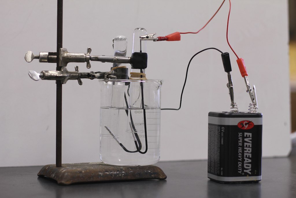 Zdjęcie przedstawia obwód elektryczny do badania przewodnictwa prądu przez roztwory wodorotlenków. dwie elektrody węglowe zanurzone w roztworze wodorotlenku w zlewce. Do elektrod przymocowane są kable, z których jeden połączony jest z żarówką a drugi ze źródłem prądu stałego (9V). Pręty połączone są z baterią stojącą na stole za pomocą kabli.
