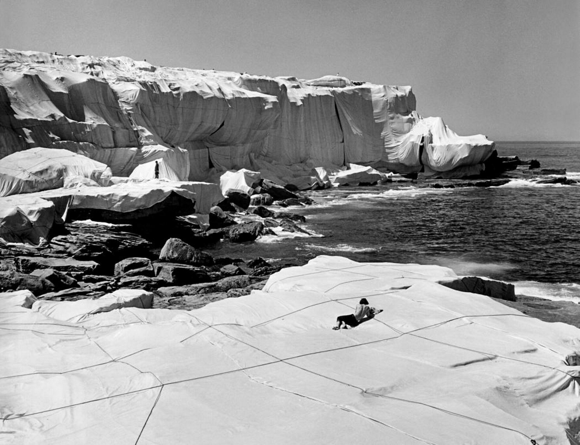Ilustracja przedstawia dzieło Christo i Jeanne-Claude. Czarno-biała fotografia ukazuje australijskie wybrzeże owinięte kwadratowymi tkaninami przymocowanymi do skał. 