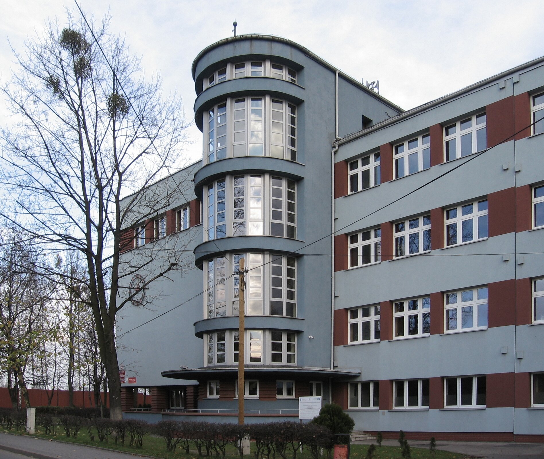 Ilustracja przedstawia budynek szkoły w Piekarach Śląskich‑Szarleju. Na zdjęciu ukazana jest frontalna część prostej budowli, w której sześcienną formę została wpisana półokrągła, oszklona, pięciokondygnacyjna bryła, wysuwająca się z fasady. Elewacja szkoły pomalowana jest na szaro, płaszczyzny pomiędzy oknami na brązowo. Budynek stoi pośród drzew.