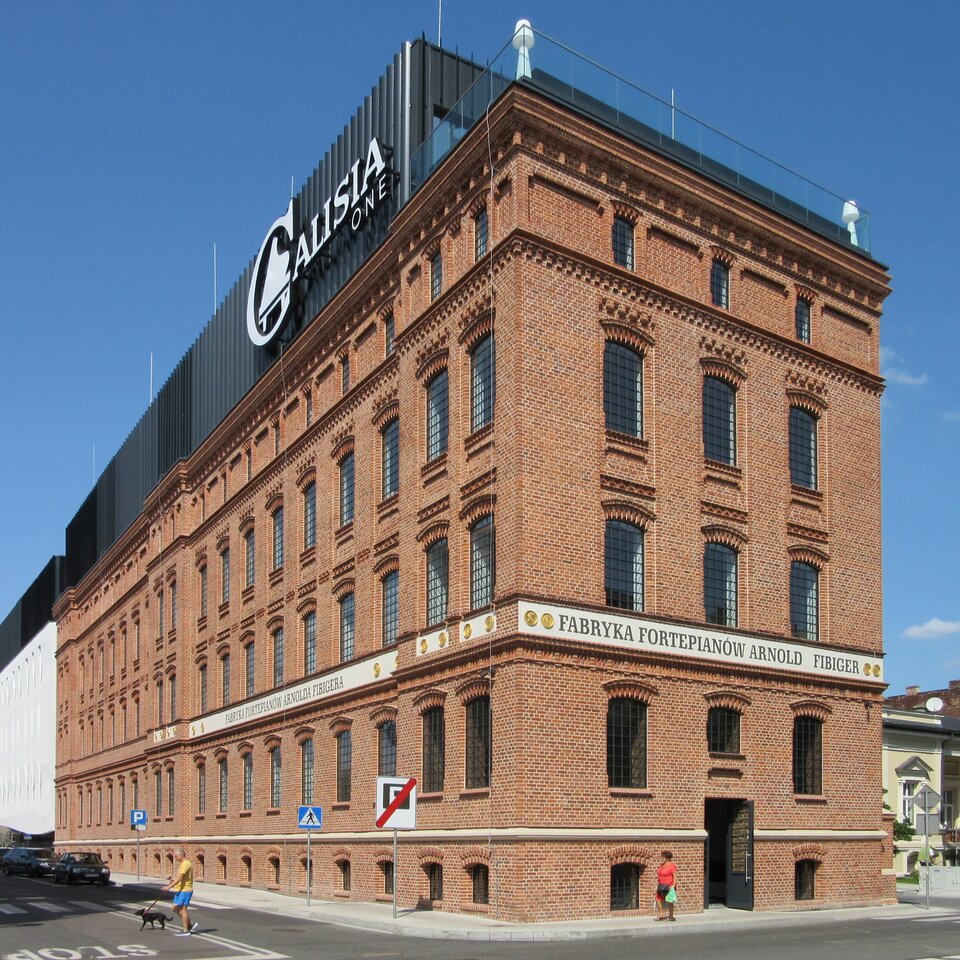 Zdjęcie przedstawia współczesny budynek kaliskiej fabryki fortepianów. Budynek z czerwonej cegły, czterokondygnacyjny. Na elewacji napis Fabryka Fortepianów Arnold Fibiger nawiązująca do historii budynku. Na współcześnie nadbudowanym elemencie dachu widnieje napis Calisia One, współczesna nazwa zakładu.