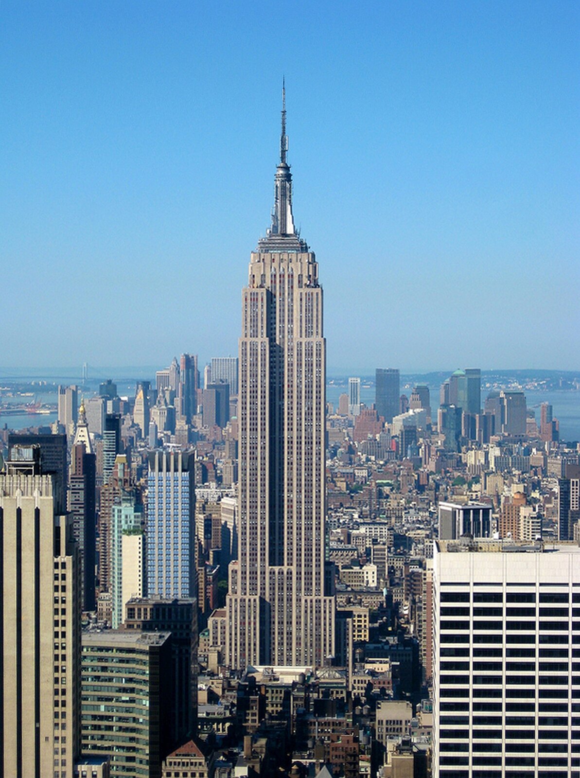 Zdjęcie przedstawia Empire State Building. Między wieżowcami i innymi budynkami miasta widać bardzo wysoki budynek z licznymi oknami oraz zwieńczony wysoką iglicą.