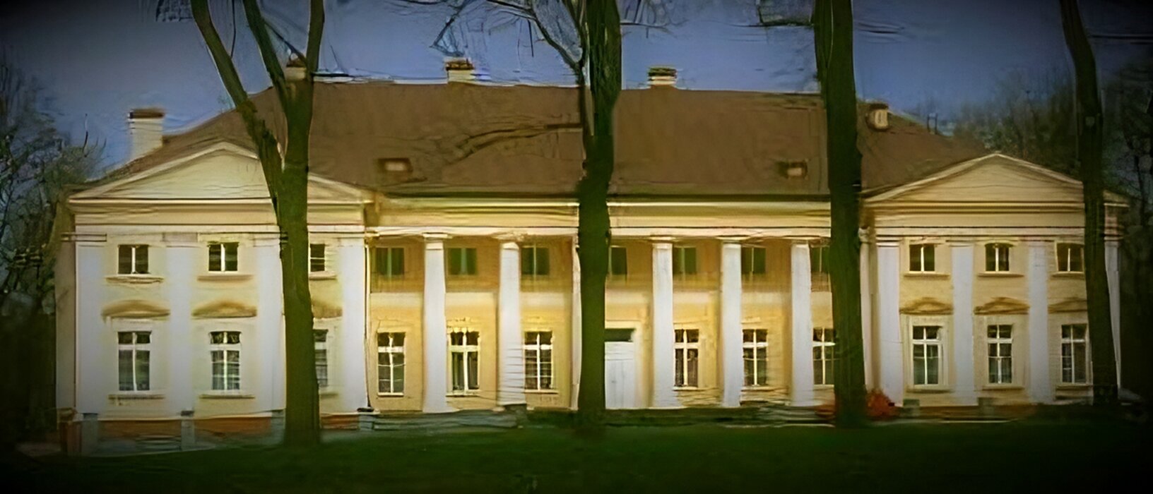 Zdjęcie przedstawia dwupiętrowy dworek, który ma spadzisty, brązowy dach. Elewacja budynku jest w kolorze żółtym, z białymi elementami ozdobnymi. W centralnej części dworku widoczne są białe, wysokie kolumny. Przed dworkiem rosną cztery gołe, wysokie, cienkie drzewa. Zdjęcie zostało wykonane w świetle dziennym.