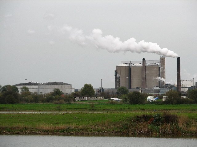 Na pierwszym planie rozciąga się pole uprawne. W tle widoczny jest rozległy zakład przemysłowy z wysokim kominem, z którego unosi się jasna smuga dymu lub pary. Zdjęcie zestawia krajobraz rolniczy z obecnością przemysłu i może ilustrować presję działalności przemysłowej na środowisko rolnicze.