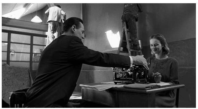 Zdjęcie przedstawia wnętrze fabryki. Robotnicy malują ściany. W sali stoi biurko, przy którym siedzi przy maszynie do pisania młoda kobieta o ciemnych włosach. Naprzeciw niej siedzi Schindler. Dotyka prawą dłonią wałka maszyny do pisania. Kobieta uśmiecha się.