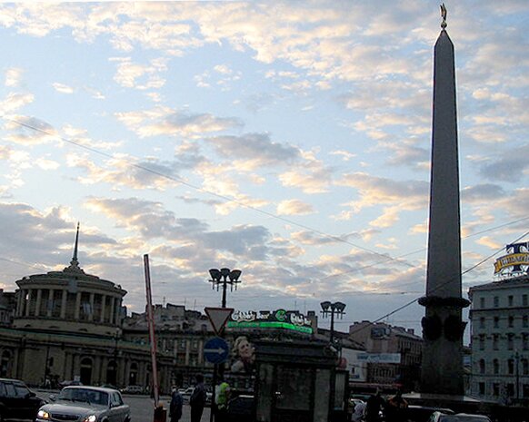 Zdjęcie przedstawia ulicę miasta. Po prawej stronie stoi pomnik, w oddali widoczne są inne budynki. W kilku miejscach stoją latarnie uliczne. Po ulicy jeżdżą samochody. Niebo nad miastem jest błękitne z niewielkim zachmurzeniem. Od chmur odbija się światło słoneczne.