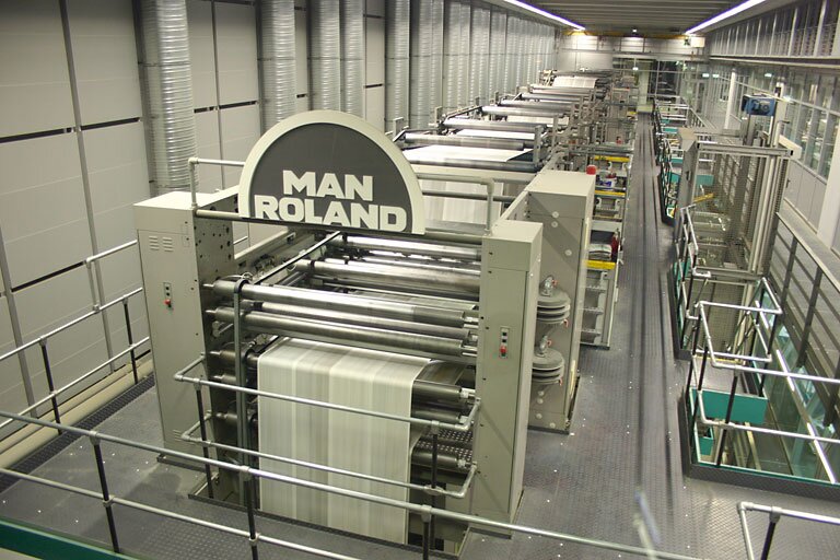 Zdjęcie przedstawia wnętrze przykładowego zakładu poligraficznego drukującego gazety. Po lewej stronie widoczne są zespoły maszyny drukującej. Na górze pierwszego zespołu widoczne jest duże półkole z napisem MAN ROLAND. Po prawej stronie rząd pojemników z farbą, schodków i elementów towarzyszących maszynie drukującej.