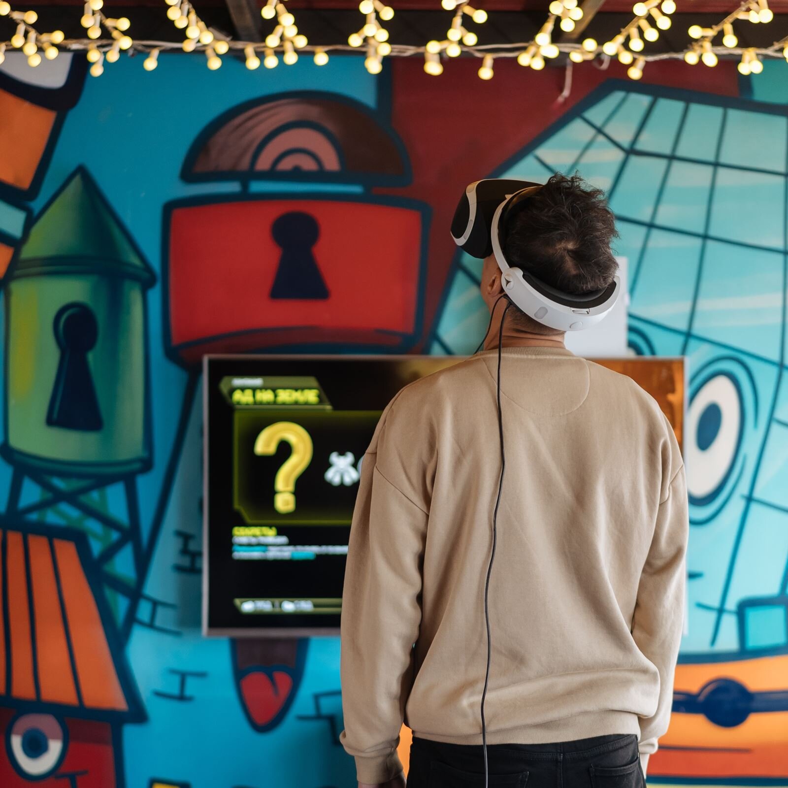 Zdjęcie przedstawia chłopca stojącego przed ekranem telewizyjnym, który umieszczony jest na tle ściany pokrytej rysunkami dziurek do kluczy. Chłopiec ma założone na głowę wirtualne gogle.